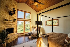 A room with a stone fireplace, large windows, a bed, a chair, and a ceiling fan. - The Ranch House (Hondo)