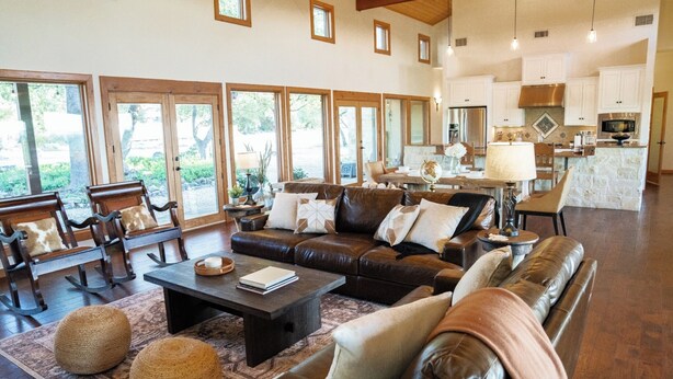 A spacious living area with leather sofas, a coffee table, and a dining table with chairs. Large windows provide natural light and views of the outdoors. - The Ranch House (Hondo)