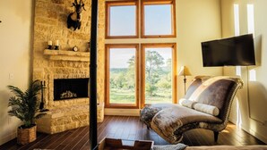 A room with a stone fireplace, a large window with a view, a leather armchair, and a flat-screen TV mounted on the wall. - The Ranch House (Hondo)