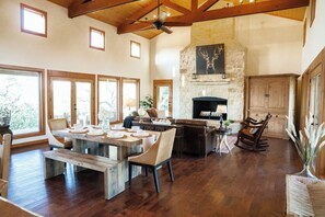 A spacious living room with a fireplace, wooden beams, and large windows. - The Ranch House (Hondo)