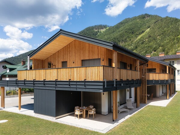 Un edificio moderno de madera de dos pisos, con un balcón, una zona de estar al aire libre techada y vistas a un paisaje montañoso.