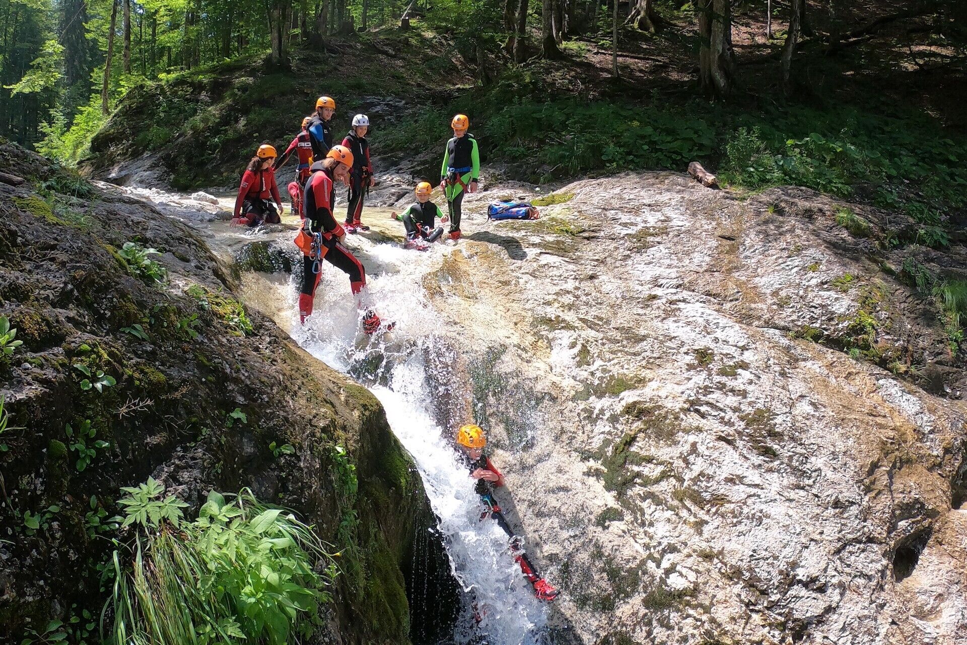 A group of people in outdoor gear near a rocky water flow in a forested area.