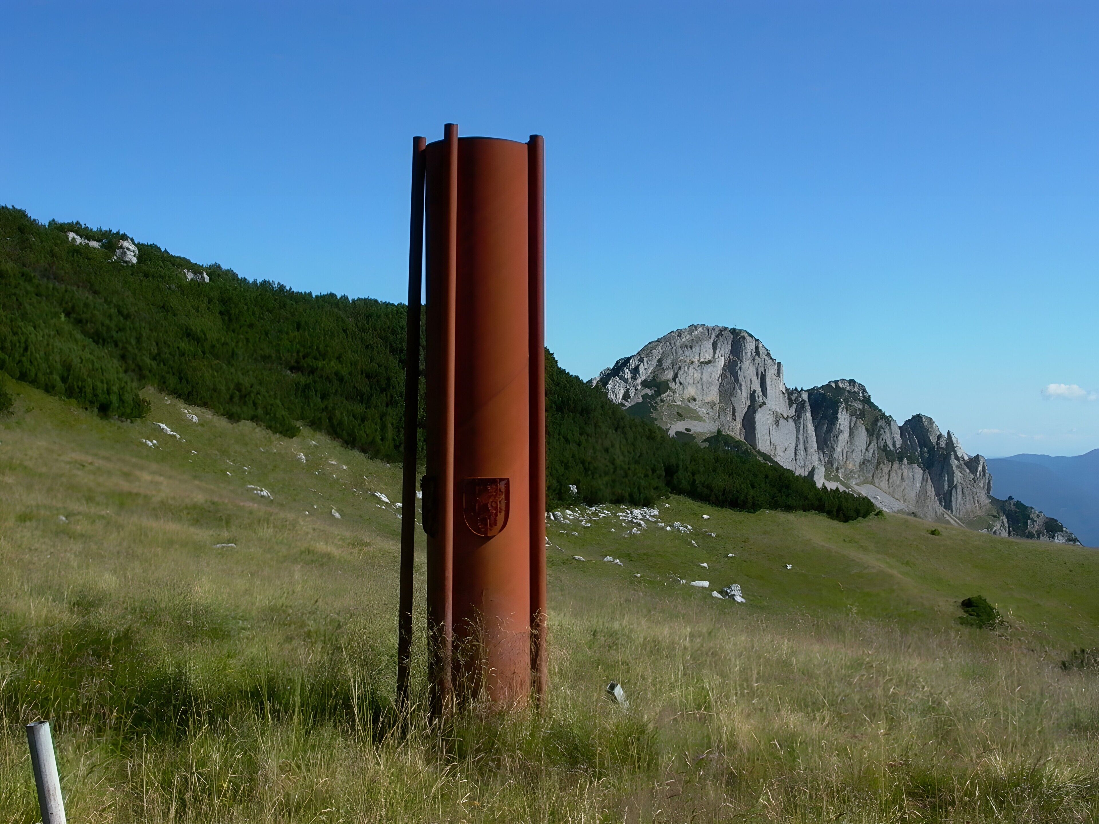 Une structure haute et cylindrique, ornée d’un emblème sur sa partie supérieure, se dressant sur un fond de paysage montagneux sous un ciel dégagé.