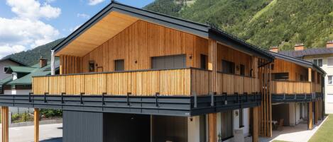 A modern two-story wooden building with a balcony, a covered outdoor seating area, and a view of a mountainous landscape.
