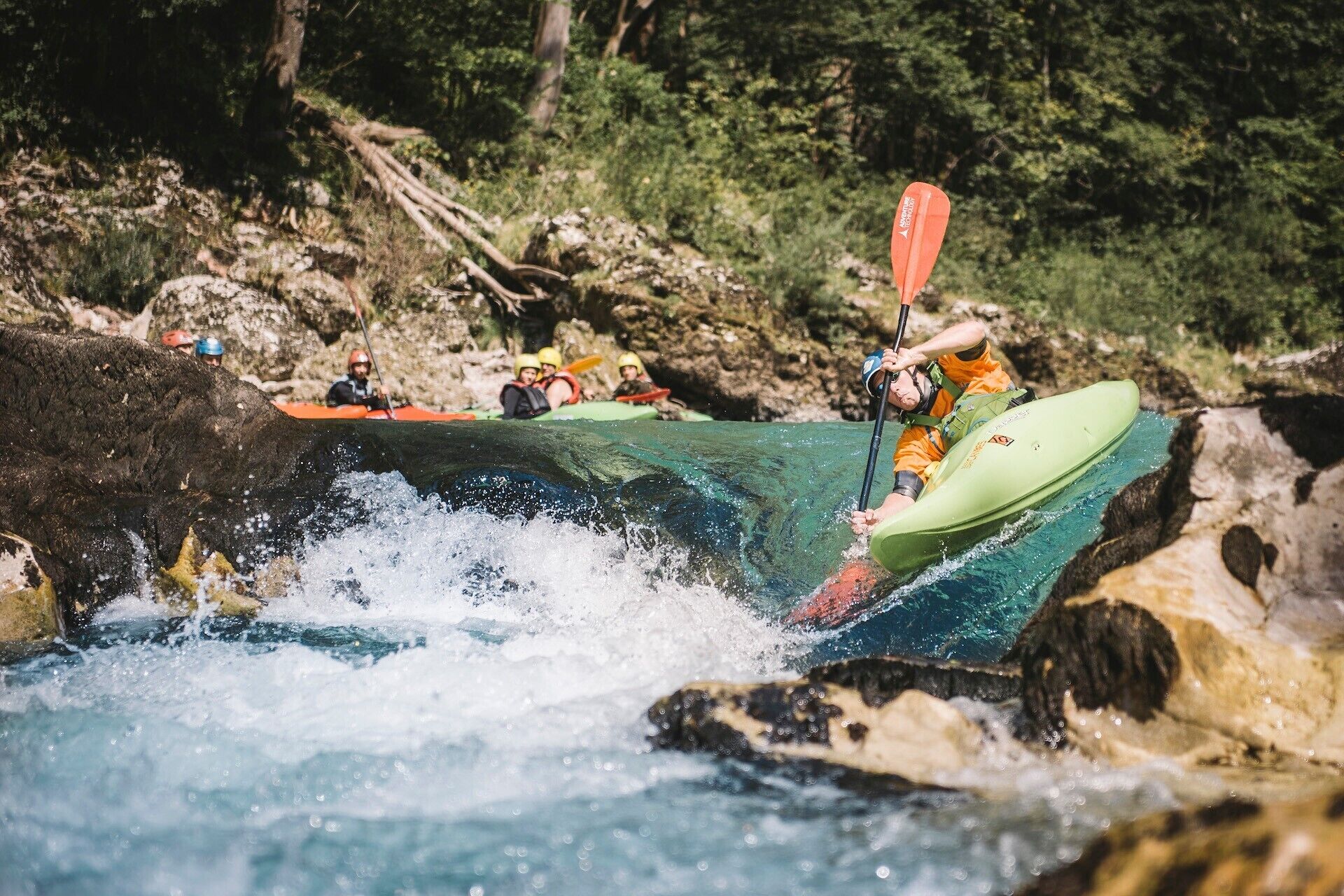 A kayaker navigating a river with a green kayak and an orange paddle.