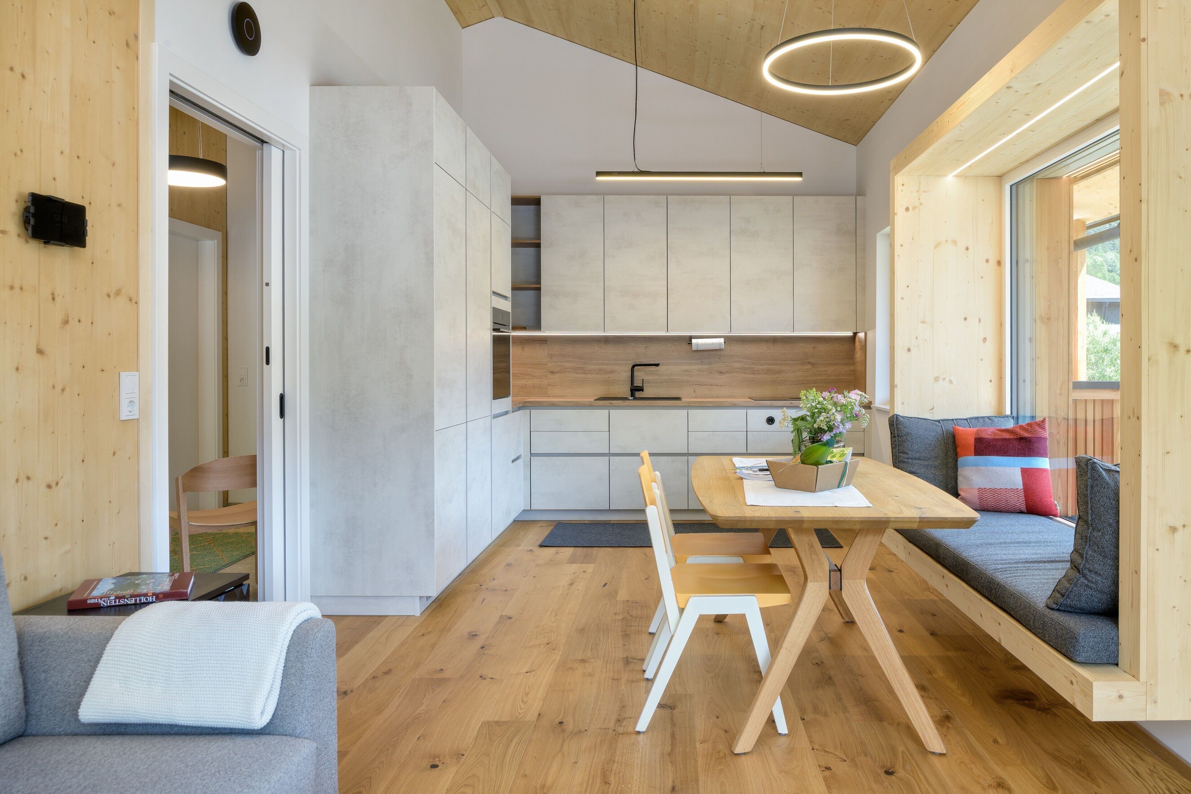 A modern kitchen with wooden cabinetry, a dining table with chairs, a built-in bench with cushions, and a view through a large window.