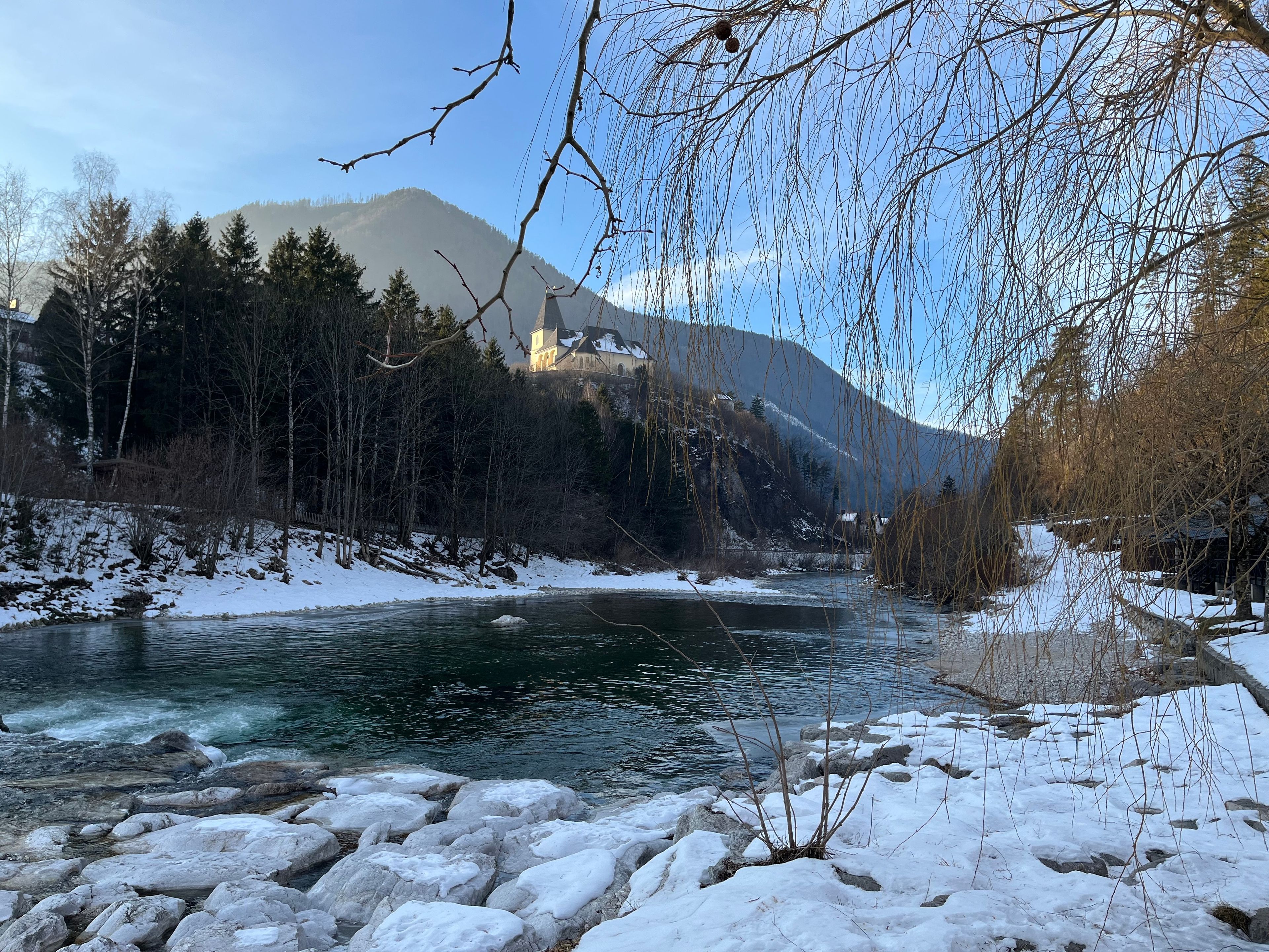 A river with snow-covered banks and a church on a hill in the background.