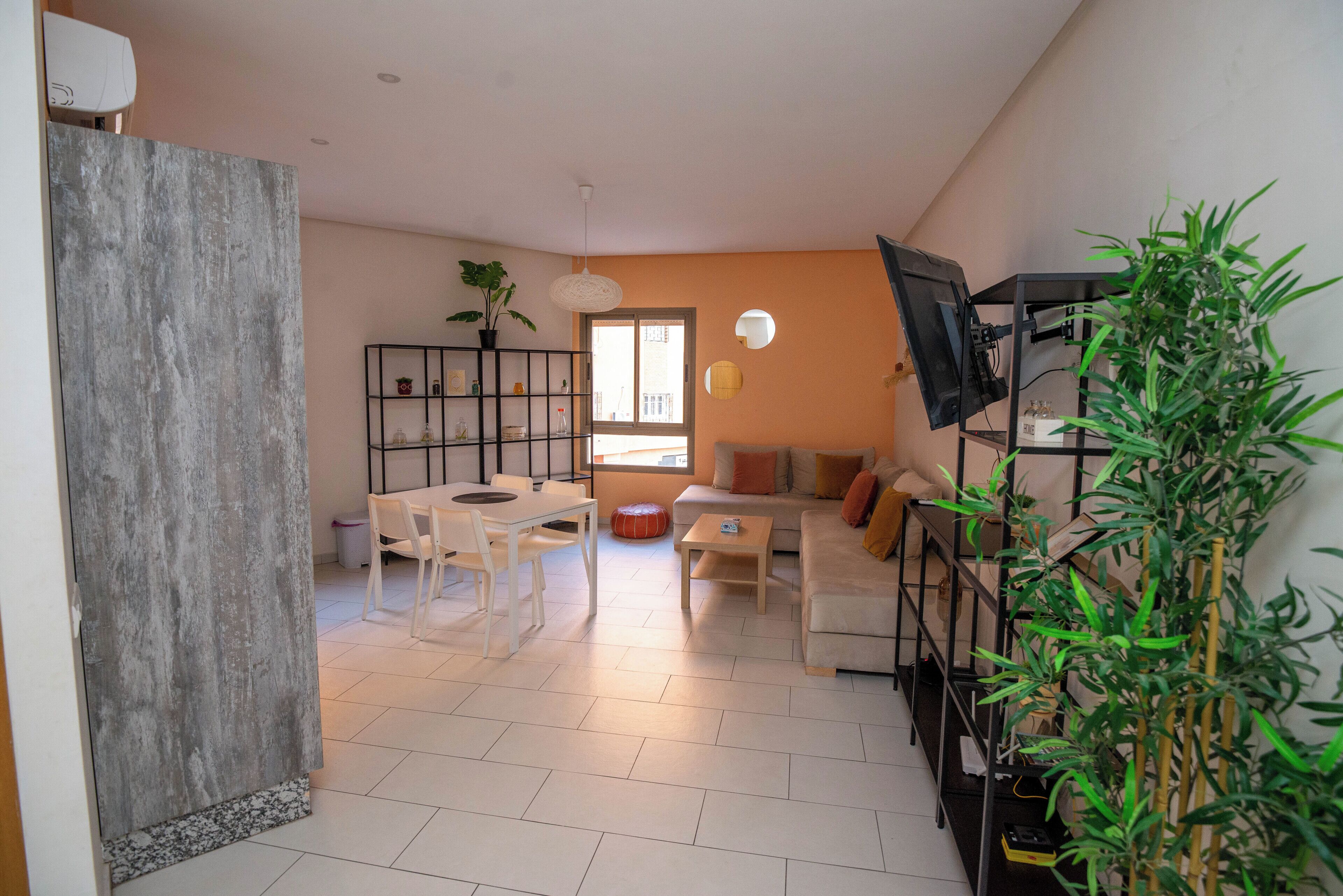 Ein Wohnzimmer mit Sofa, Couchtisch und Esstisch mit Stühlen. An der Wand ist ein Fernseher angebracht und in der Ecke steht eine Pflanze.