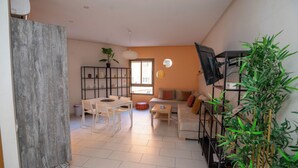 Ein Wohnzimmer mit Sofa, Couchtisch und Esstisch mit Stühlen. An der Wand ist ein Fernseher angebracht und in der Ecke steht eine Pflanze.