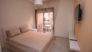 Ein Schlafzimmer mit Bett, Nachttisch, Fernseher, einem Fenster mit durchsichtigen Vorhängen und Blick auf einen Außenbalkon.