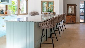 A kitchen with a central island, bar stools, a wooden cabinet, and a view through the window. - Exclusive Mediterranean Villa with Private Pool in Cala d'Or (Santanyí)