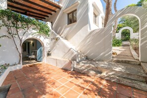 Um edifício branco com um pátio ladrilhado, uma pérgula de madeira e uma escadaria que leva a um andar superior.