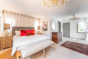 Quarto com cama grande, mesas de cabeceira, penteadeira, um banco, lustre e vista para o exterior.