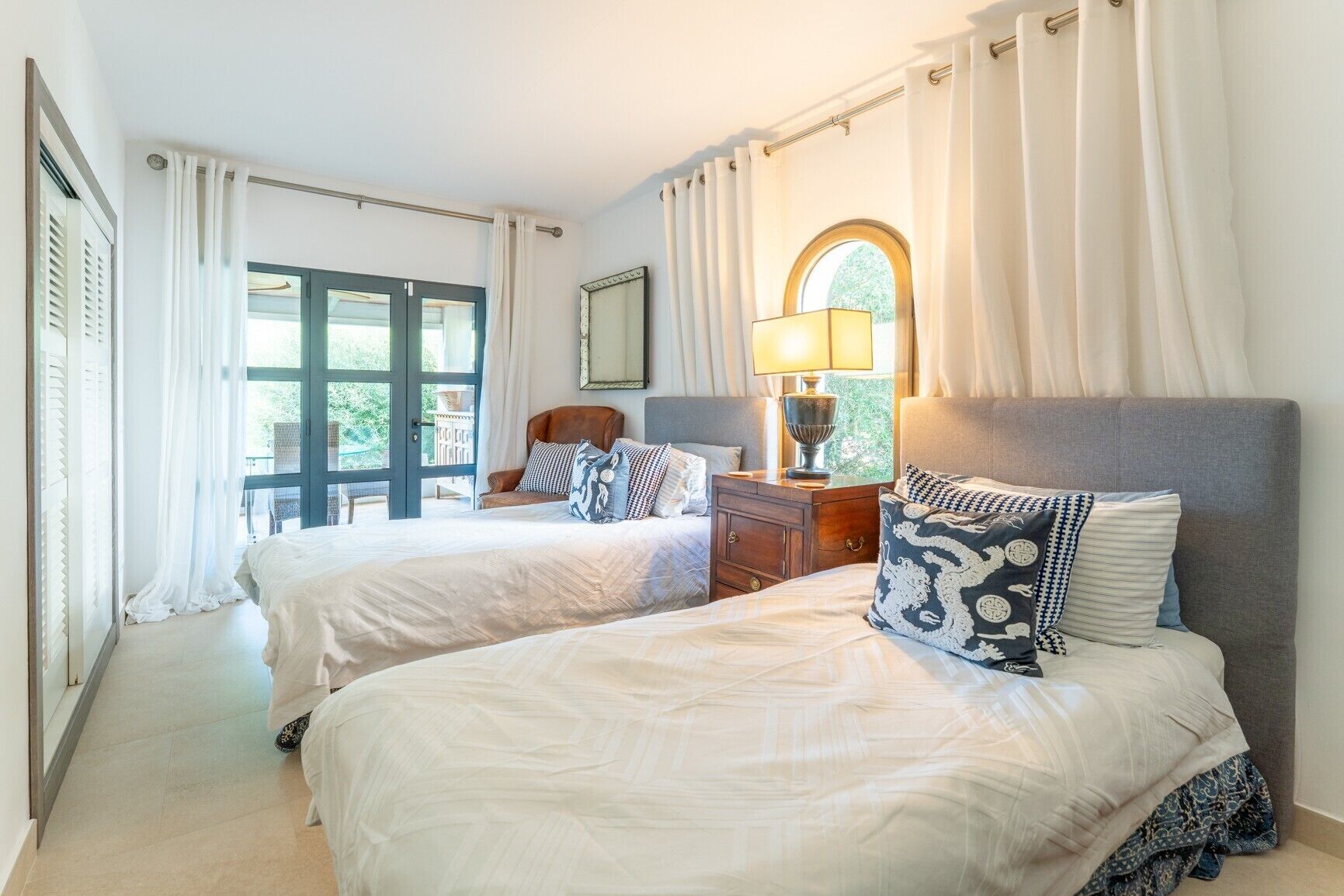A hotel room with two beds, a window with curtains, a lamp, and a view of the outside.