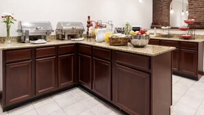 Una cocina con encimera de granito, armarios de madera oscura, pared de ladrillo, un reloj y diversos alimentos.