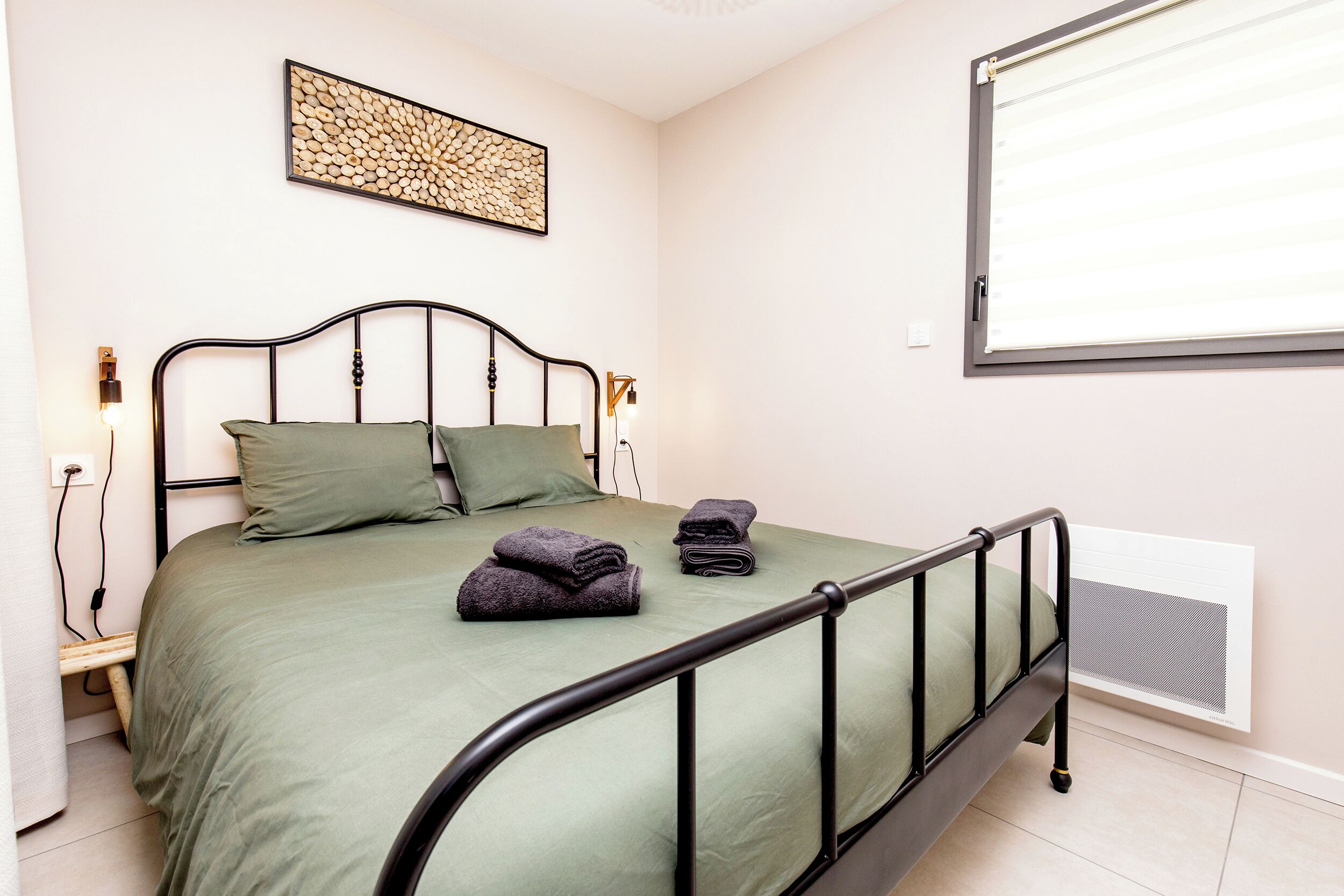 Un lit simple avec un cadre en métal, une couverture verte, deux oreillers et une serviette pliée posée sur le lit.