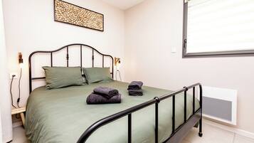 Un lit simple avec un cadre en métal, une couverture verte, deux oreillers et une serviette pliée posée sur le lit.