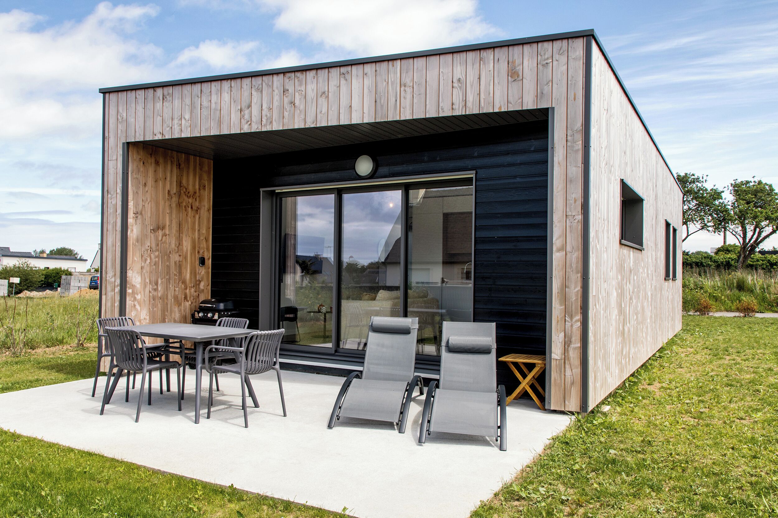 Ein modernes Haus mit einer großen Schiebeglastür, einer Terrasse mit Tisch und Stühlen sowie einer kleinen Holzterrasse.