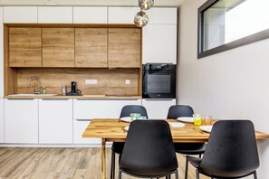 A modern kitchen with wooden cabinets, a black microwave, and a dining area with black chairs and a wooden table. - TI KAZ BREIZH, a new wooden house (Cléder)