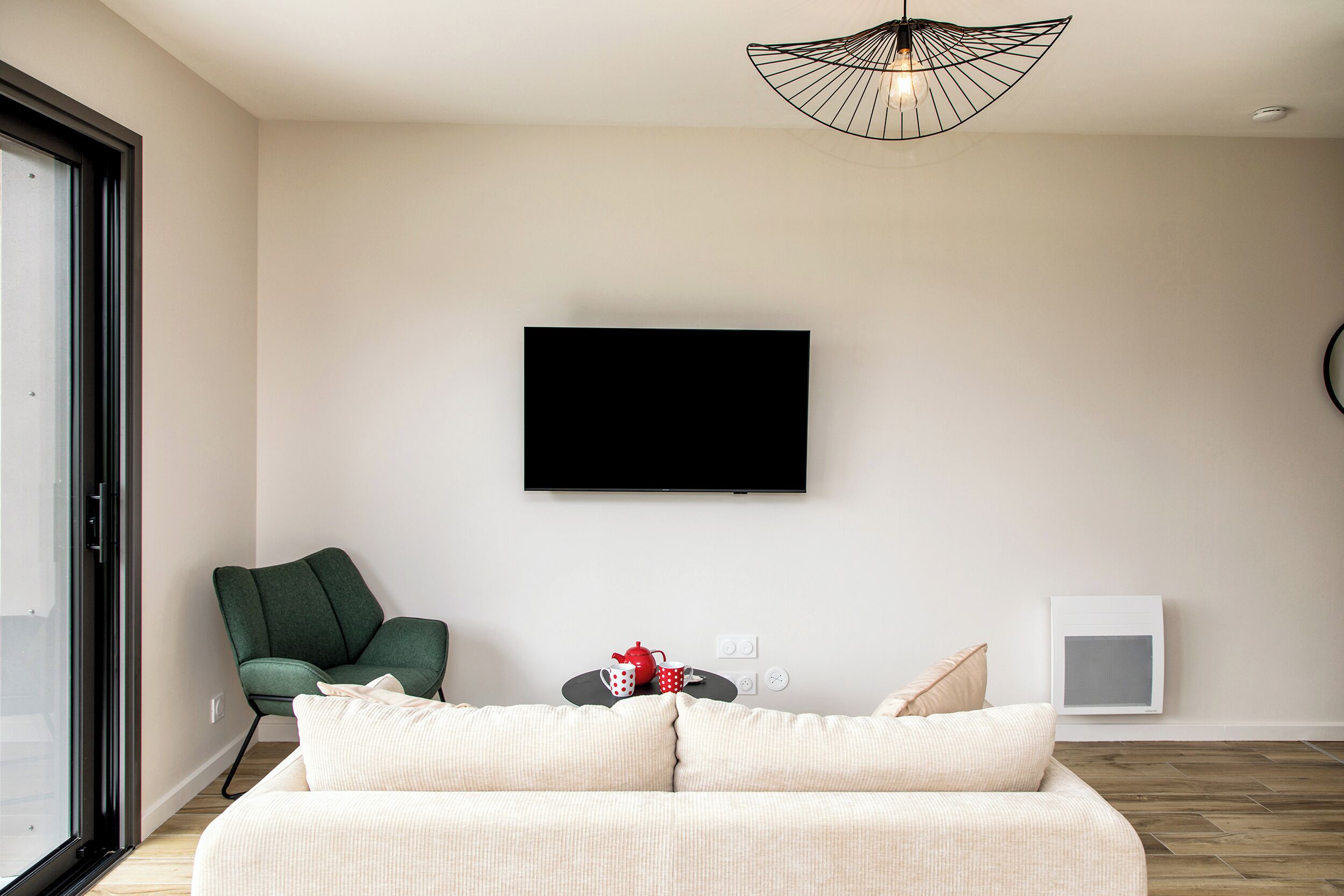 Un salon moderne comprenant un canapé, un fauteuil vert, un téléviseur à écran plat, une petite table agrémentée d’objets décoratifs et une fenêtre offrant une vue agréable.