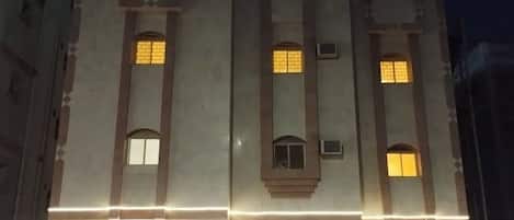 Un immeuble à plusieurs étages, doté d’un escalier central, de nombreuses fenêtres et d’une voiture garée devant.