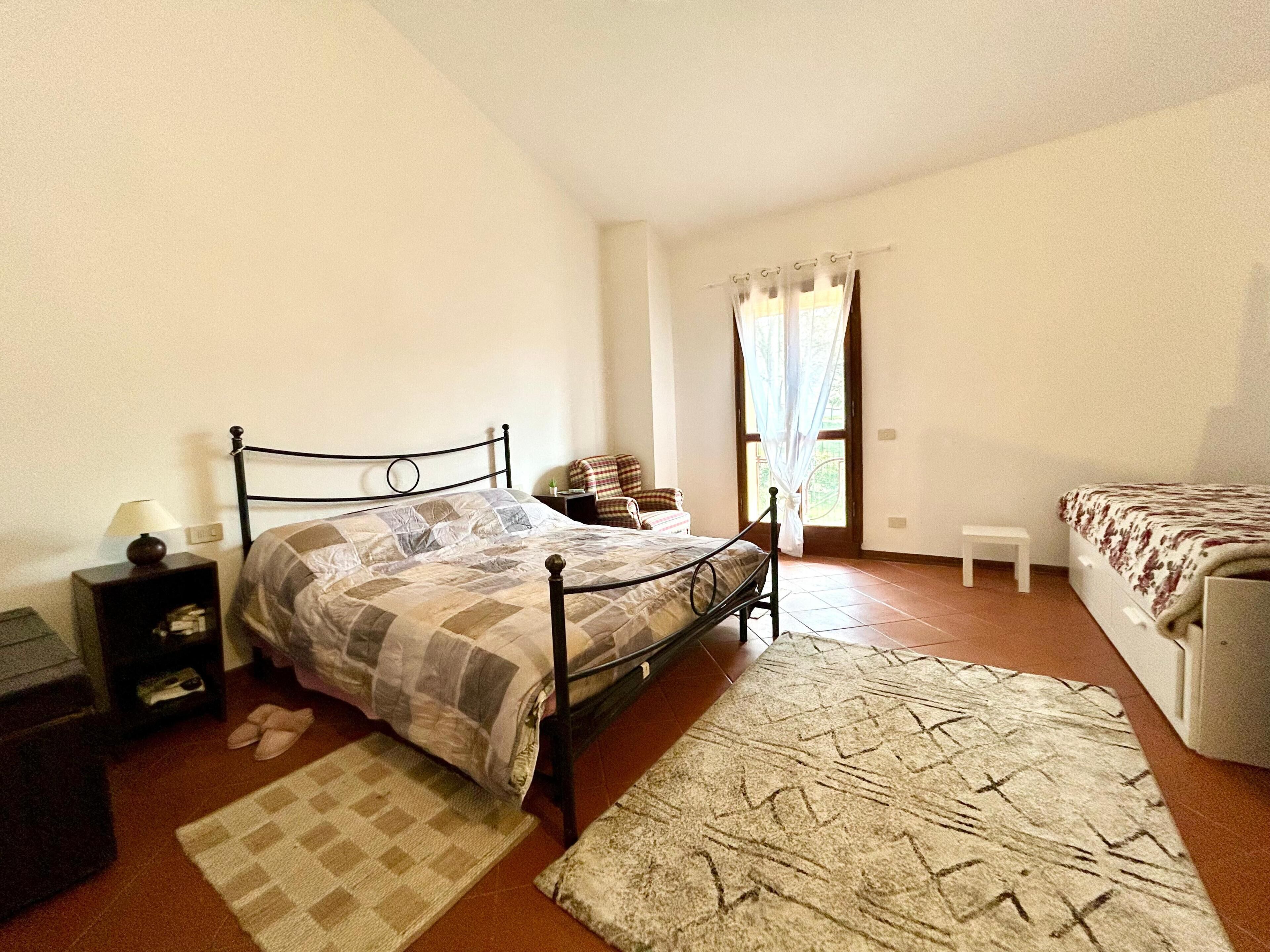 Ein Schlafzimmer mit Bett, Nachttisch, Lampe, Stuhl, Fenster und einem kleinen Hocker.