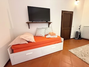 A bed with an orange mattress, a TV mounted above it, a white bedside table with drawers, a door, and a vacuum cleaner.