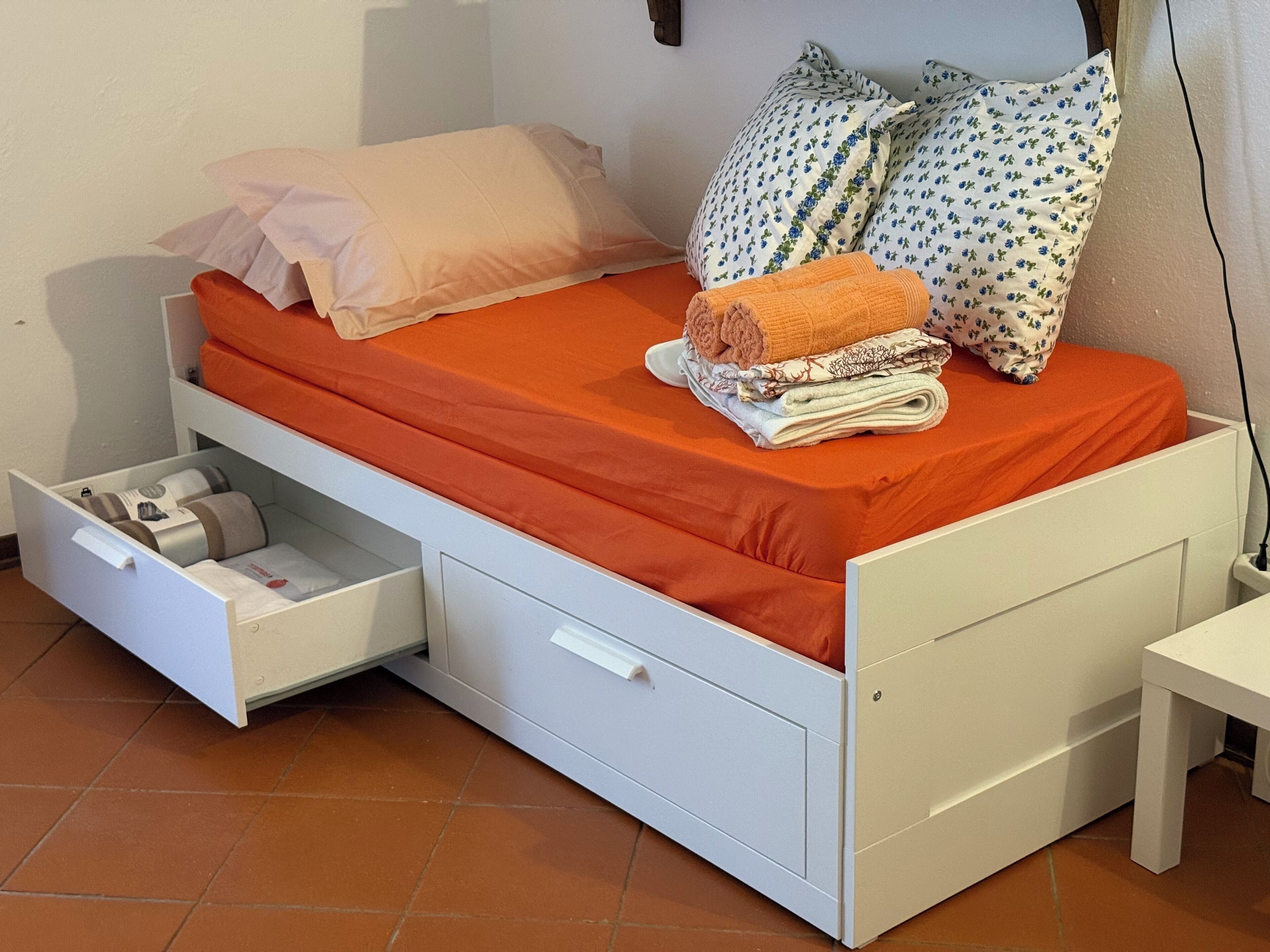 Habitación individual con una colcha naranja, almohadas blancas, un cajón lateral y una mesita de noche pequeña.