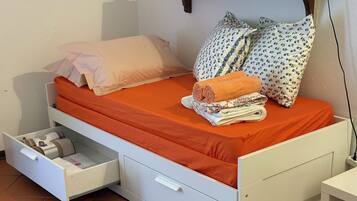 Habitación individual con una colcha naranja, almohadas blancas, un cajón lateral y una mesita de noche pequeña.