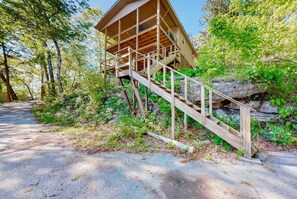 Eine Holzhütte mit einer Treppe, die zu einer Terrasse führt, umgeben von Bäumen und einem Kiesweg.