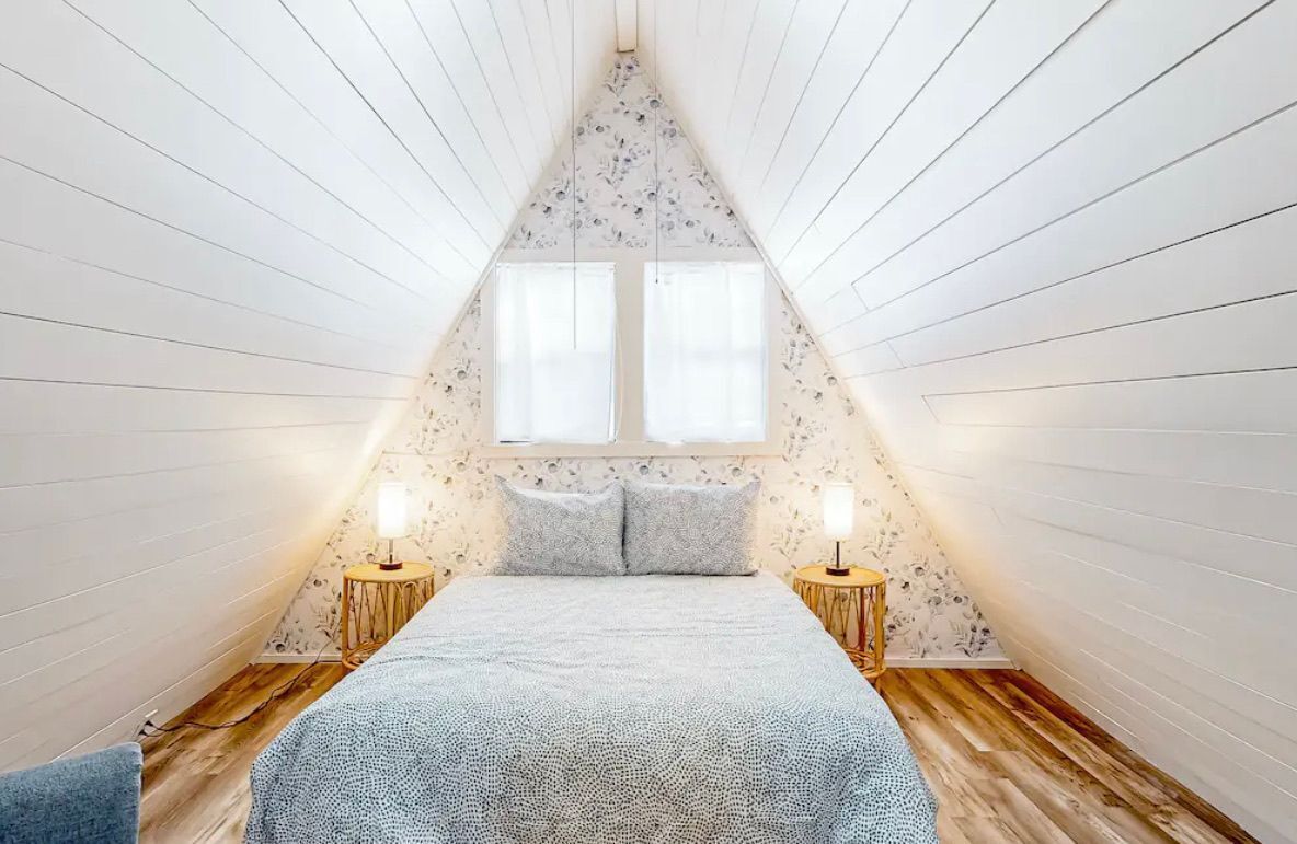 Un dormitorio con techo inclinado, dos ventanas, una cama con colcha estampada, dos lámparas de noche y suelo de madera.