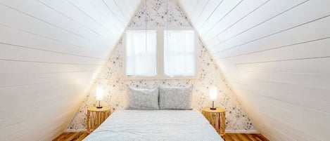 Quarto com teto inclinado, duas janelas, cama com colcha estampada, duas luminárias de cabeceira e piso de madeira.