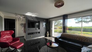 Ein modernes Wohnzimmer mit einem Flachbildfernseher, einem roten Ledersessel, einem schwarzen Sofa und einem großen Fenster mit Aussicht.