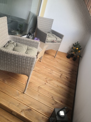 A wicker chair with cushions, a small table, and a potted plant on a wooden deck. - Vacation rental overlooking the lake in Bad Weißenstadt, with a view of the lake-side boarding house  (Weißenstadt)