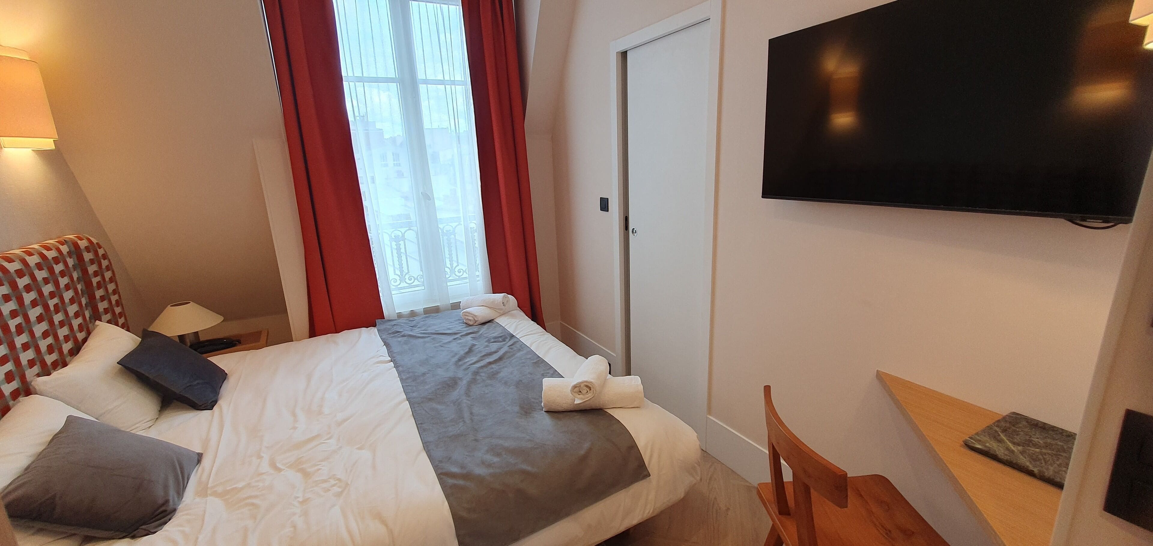 Una habitación de hotel con cama, televisión, ventana con cortinas, mesita de noche y una silla.