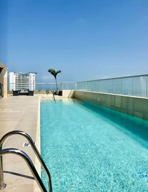 A rooftop pool with clear blue water, a glass railing, and a palm tree. - Ivory Tide Coastal Apt • Ocean View • Beach WiFi (Cartagena)