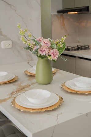 A dining table with a green vase of flowers, white plates with gold rims, and a marble countertop. - Ivory Tide Coastal Apt • Ocean View • Beach WiFi (Cartagena)
