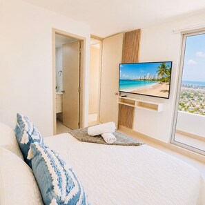 A hotel room with a bed, a television displaying a beach scene, a window with a city view, and a door leading to another room. - Ivory Tide Coastal Apt • Ocean View • Beach WiFi (Cartagena)