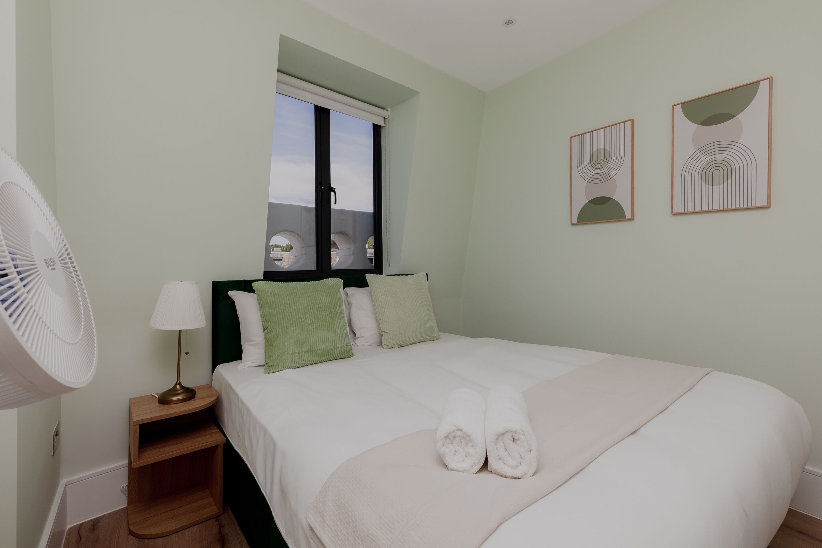 A neatly made bed with two rolled towels, a bedside table with a lamp, a standing fan, and two framed artworks on the wall.