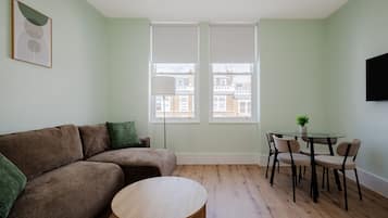 Een woonkamer met een bruine bank, een ronde houten tafel en een eettafel met glazen blad en stoelen.