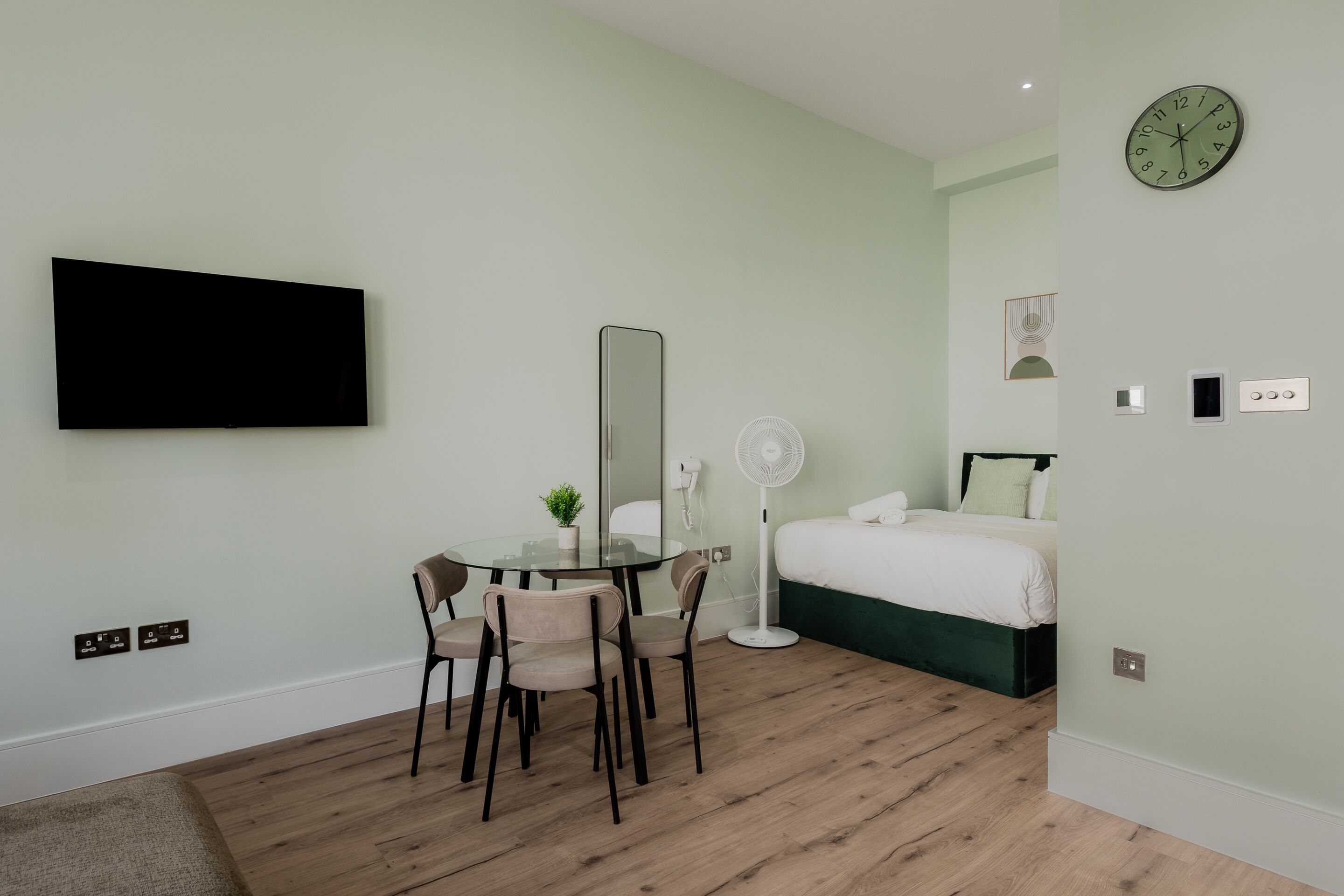 Habitación de hotel con cama, escritorio con sillas, televisor, espejo, reloj y ventilador.