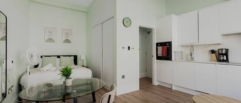 Een moderne hotelkamer met een glazen eettafel, een bed met groene beddengoed, een kleine plant en een keuken met witte kasten en een klok.