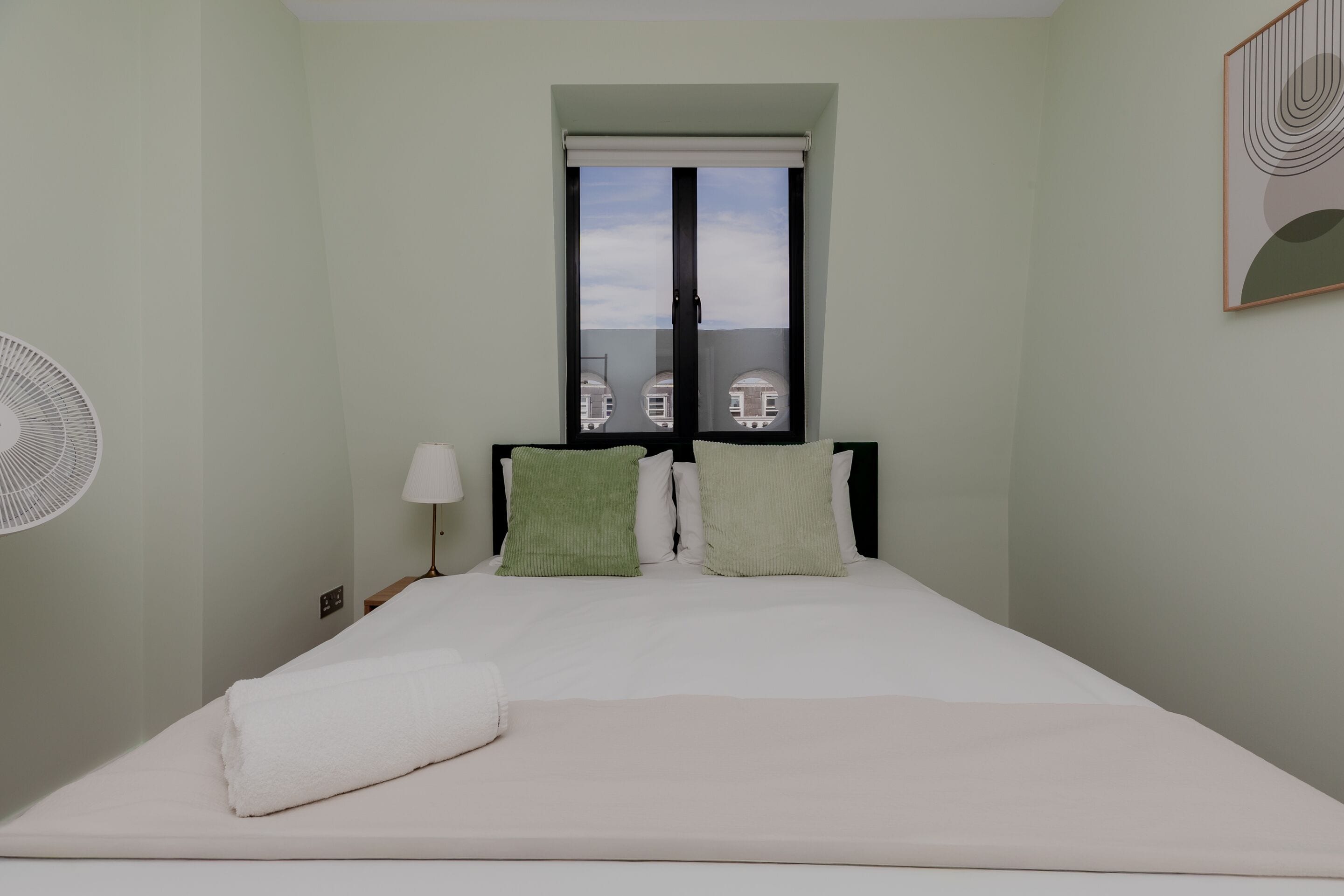 Een slaapkamer met een bed, een raam met jaloezieën, een bedlamp en een ventilator aan de muur.
