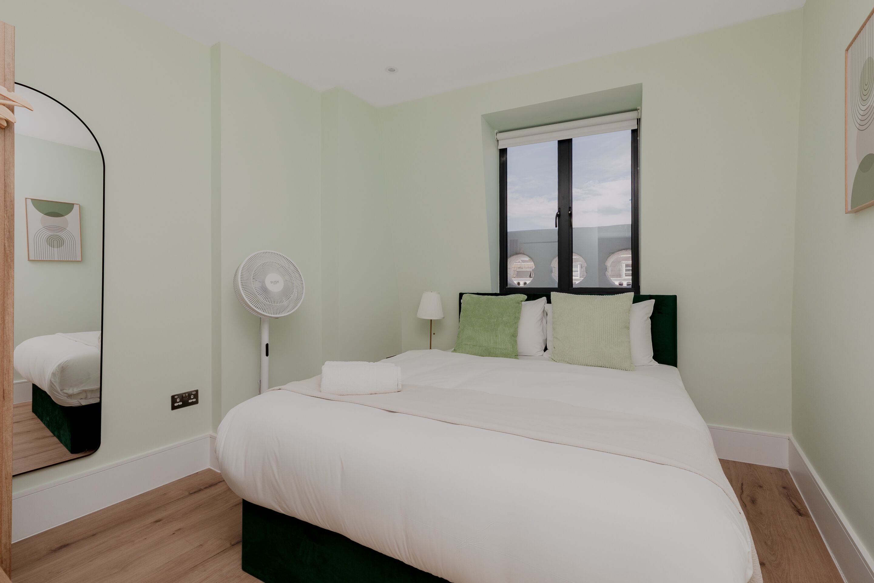 A bedroom with a bed, a standing fan, a window, and a mirror.