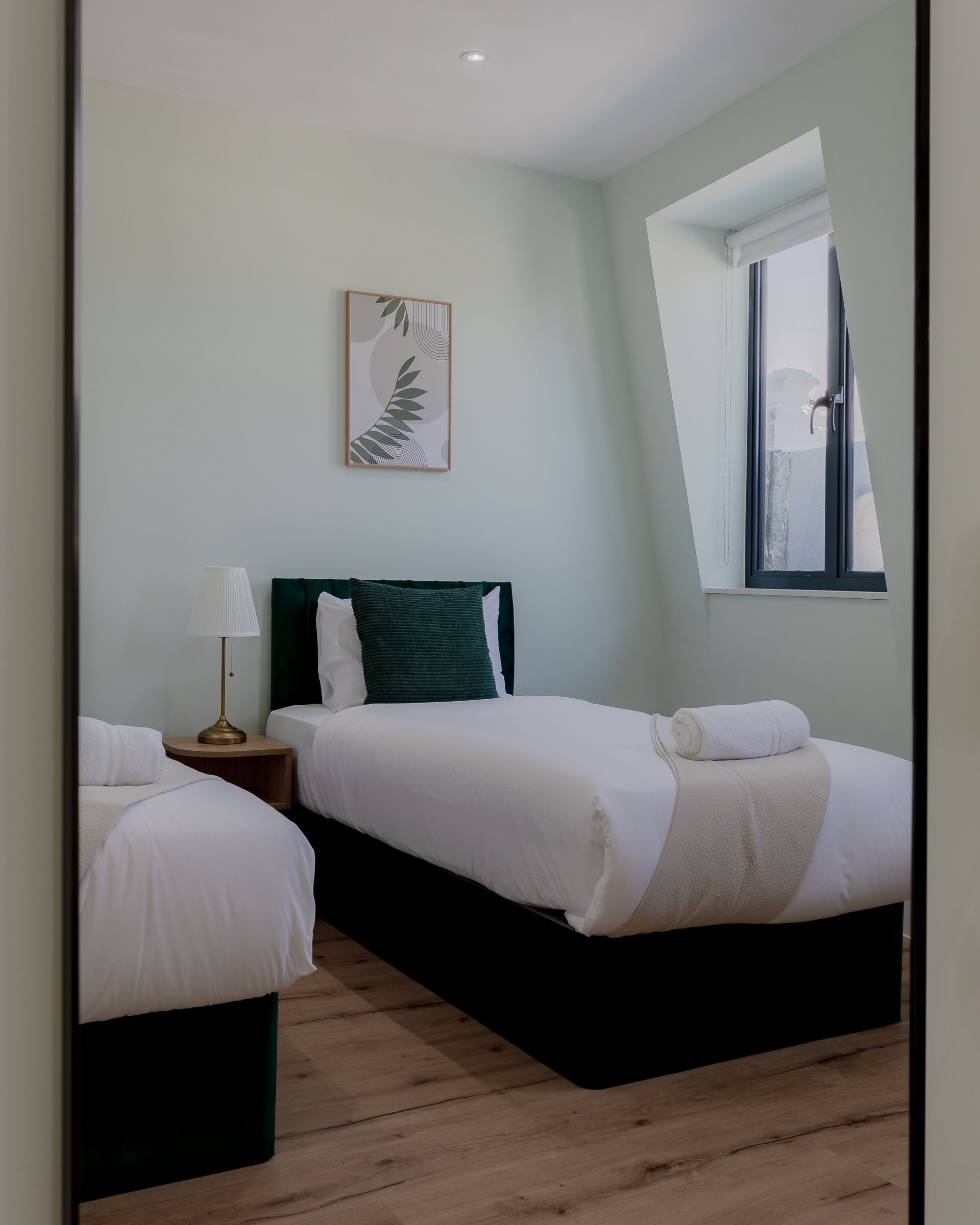 A bedroom with two beds, a nightstand with a lamp, a framed artwork on the wall, and a window with a view.