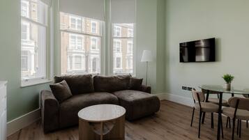 A modern living room with a sectional sofa, a round coffee table, a dining table with chairs, and a flat-screen TV mounted on the wall.
