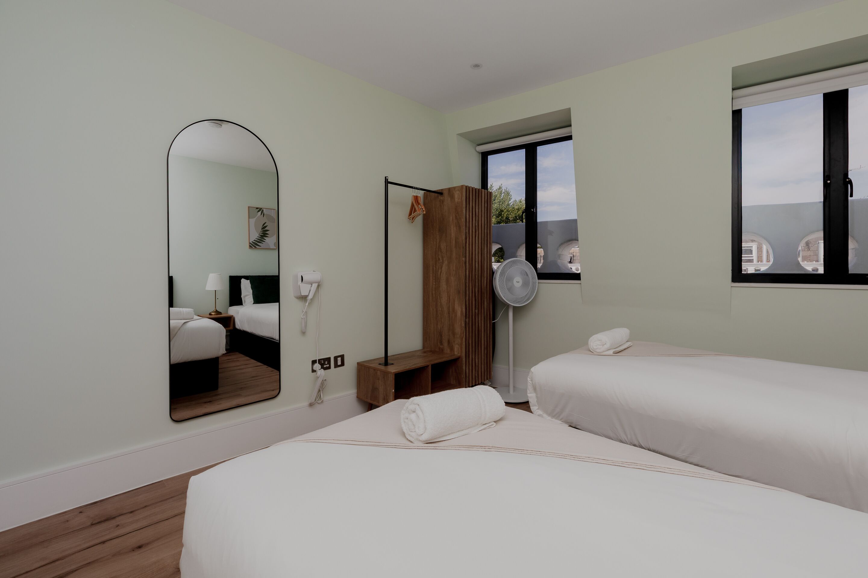 Habitación de hotel con dos camas, un espejo grande, un armario de madera, una mesita de noche, un ventilador y una ventana con vistas.