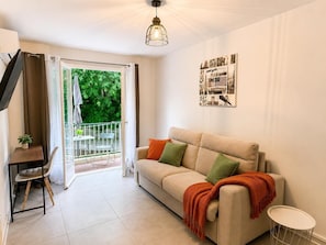 Un salon comprenant un canapé, une petite table, un fauteuil, un balcon donnant sur un espace verdoyant et un luminaire au plafond.