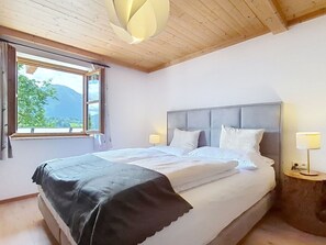 A bedroom with a large bed, a wooden ceiling, a window with a view of greenery, and a nightstand with a lamp.
