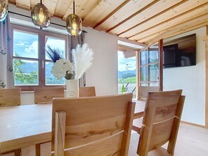 A dining area with wooden tables and chairs, a vase with white flowers, and large windows with a mountain view.